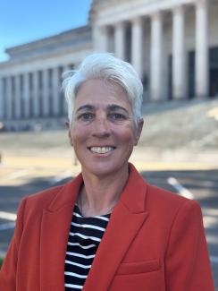 A smiling Jo Aiken stands in a red suit jacket with the Legislative Building's North steps in the background.