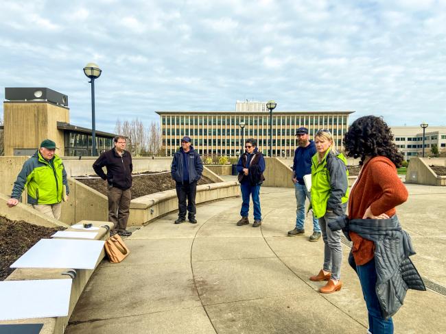 Seven staff members from DES and Woodland Park Zoo stand in a semicircle on the grounds of the future garden, discussing the design plans