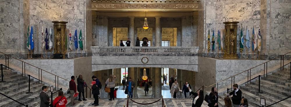 Legislative Building staff and visitors walking through the Rotunda. Some people are talking to each other, while others take pictures of the building's marble and brass features.