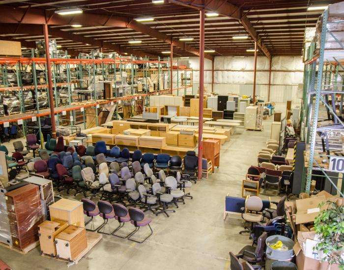 A high-angle view of various office equipment, including rolling chairs, cabinets, and desks, organized on a well-lit warehouse floor.