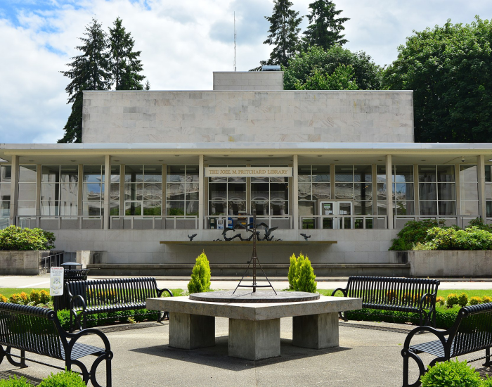 A view of the Joel Pritchard State Library's front side, which is mainly made of white stone with large glass walls. A fountain, a sundial, four metal park benches, and various green bushes are in front of the building.