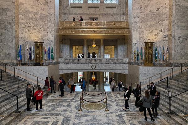 Legislative Building staff and visitors walking through the Rotunda. Some people are talking to each other, while others take pictures of the building's marble and brass features.